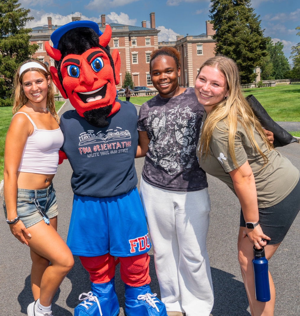 college students stand and smile with the FDU Ian the Devil mascot. The Hennessy Hall mansion stands behind them.