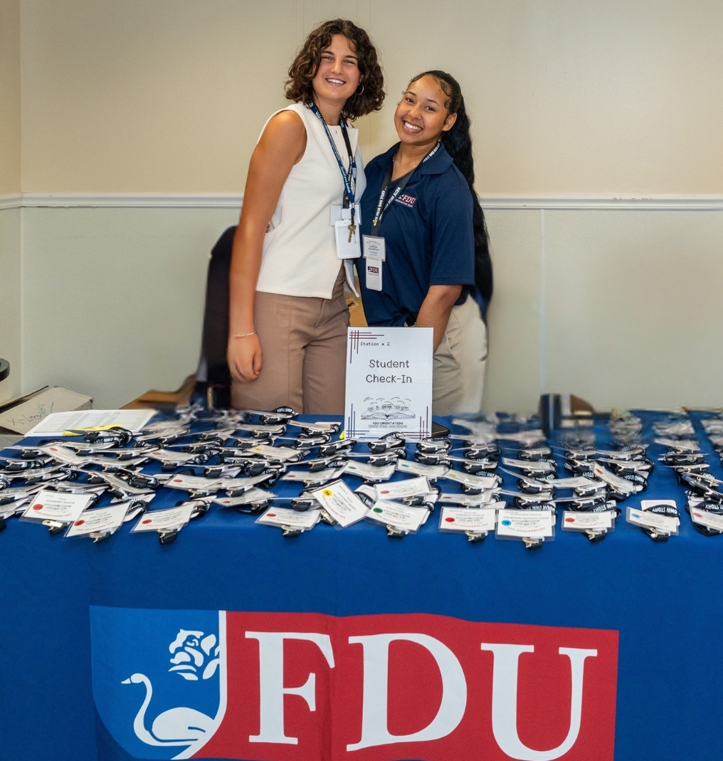 two students smile for the camera. they stand at a table for student check in. the table is covered in name tags and a tablecloth with the FDU logo.