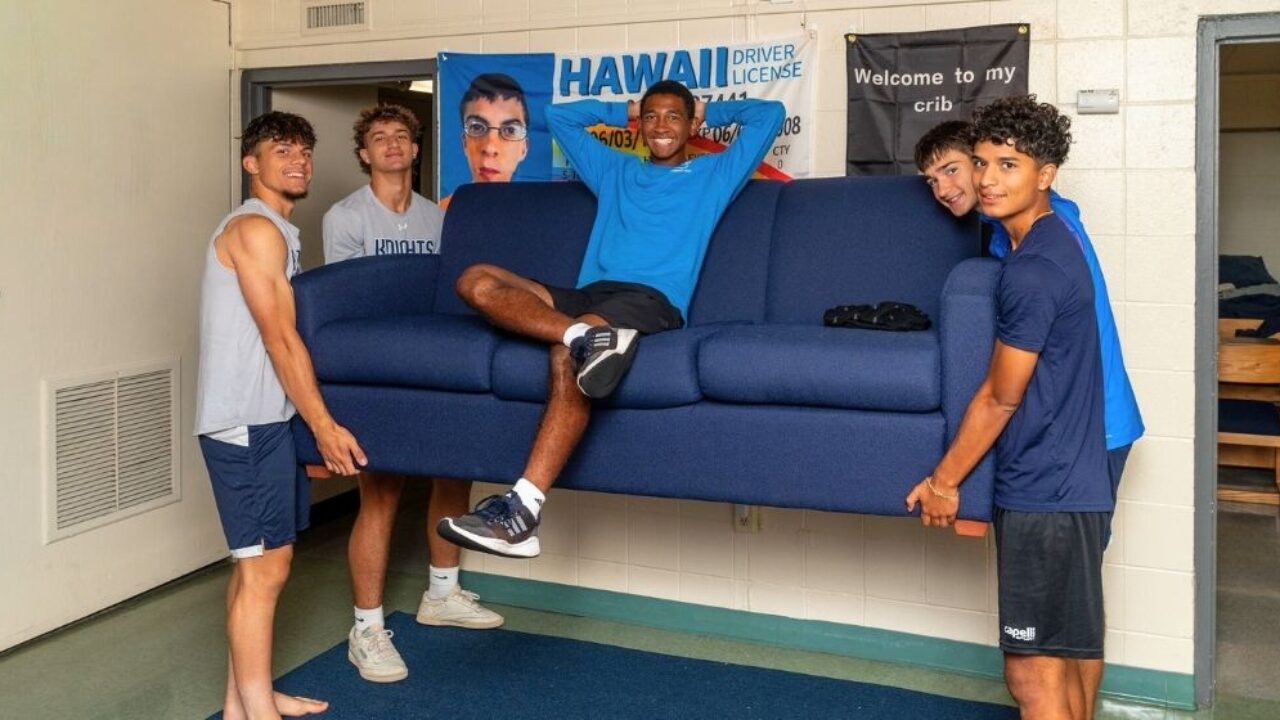 a group of students lift up a couch. another student still sits on the elevated couch, with his arms crossed behind his head.