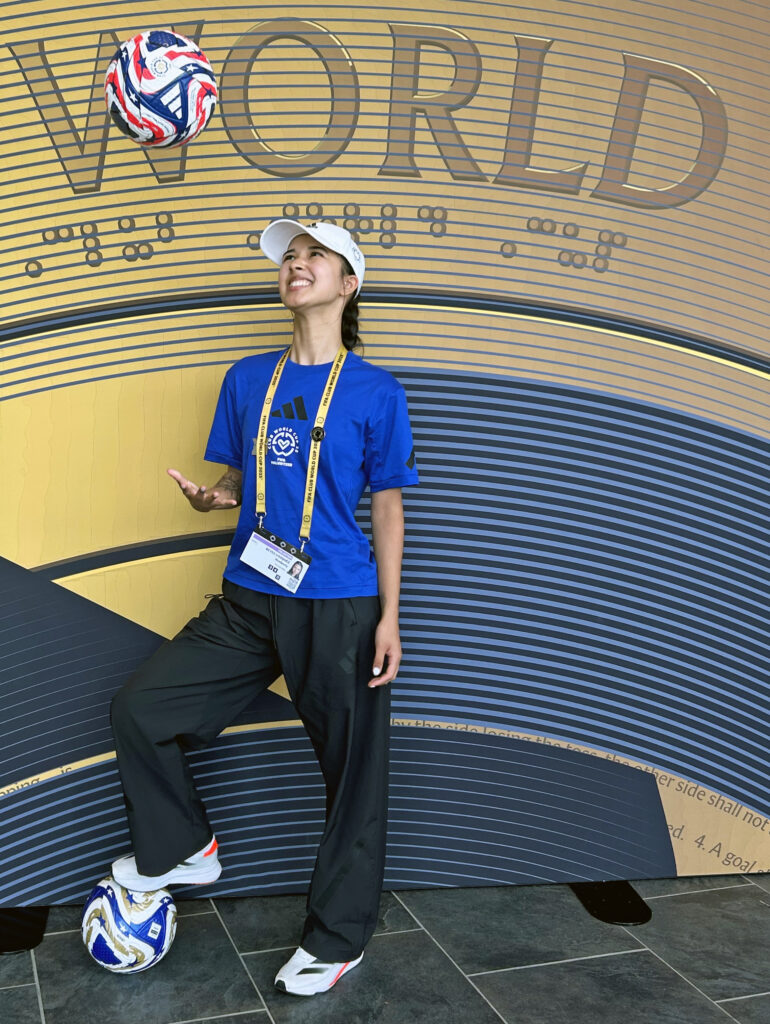 A young woman in a FIFA volunteer uniform throws a soccer ball in the air.