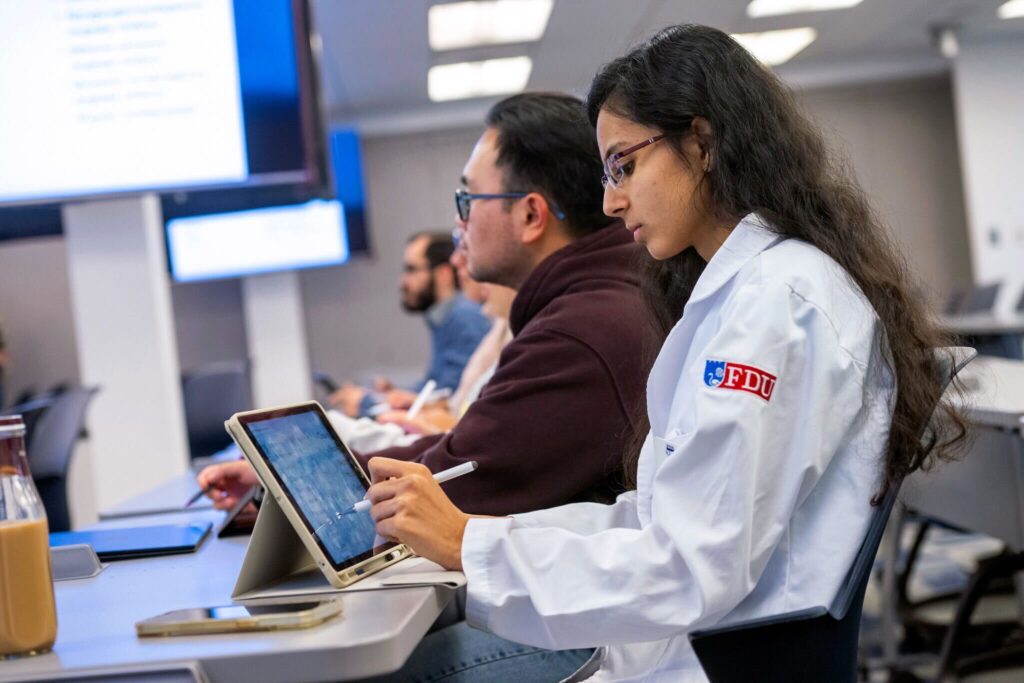 students sit and look up to the front of a classroom. one student wears a white lab coat with the FDU logo on the coat's arm. she reviews her notes on an iPad.