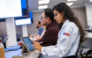 students sit and look up to the front of a classroom. one student wears a white lab coat with the FDU logo on the coat's arm. she reviews her notes on an iPad.