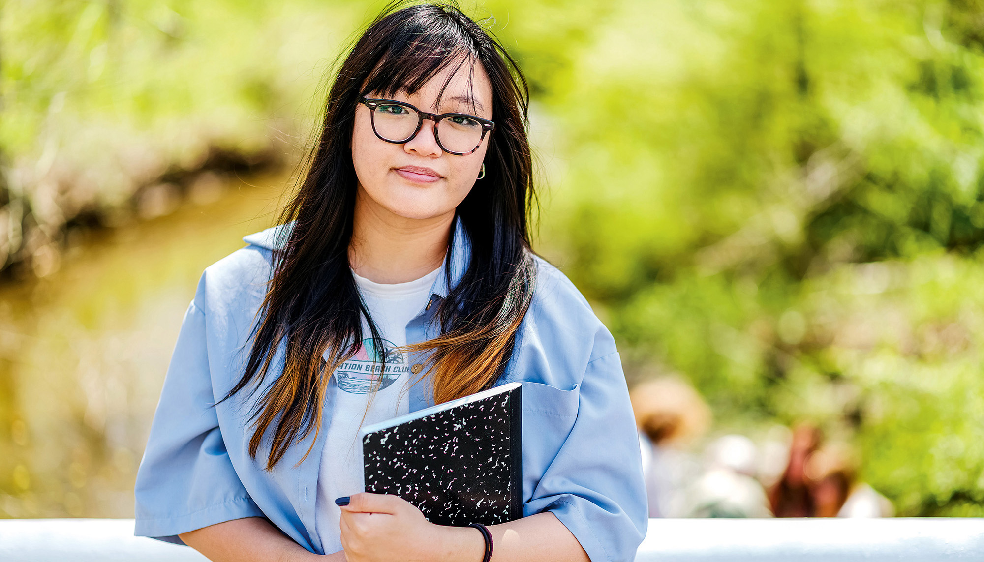 A young woman wearing glasses holds a notebook while out doing fieldwork. A stream and trees are in the background.