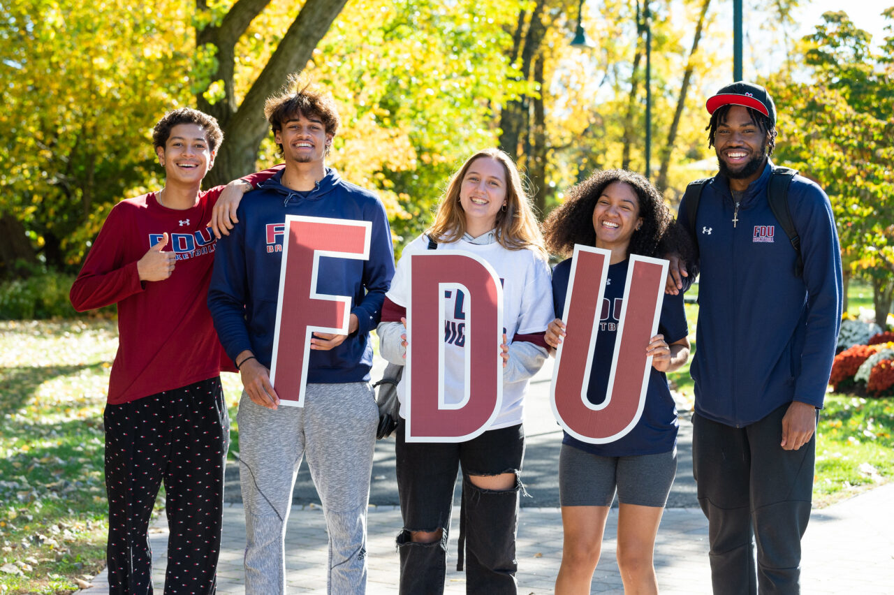 A group of male and female students stand together outside on the Metropolitan Campus, holding up FDU letters.