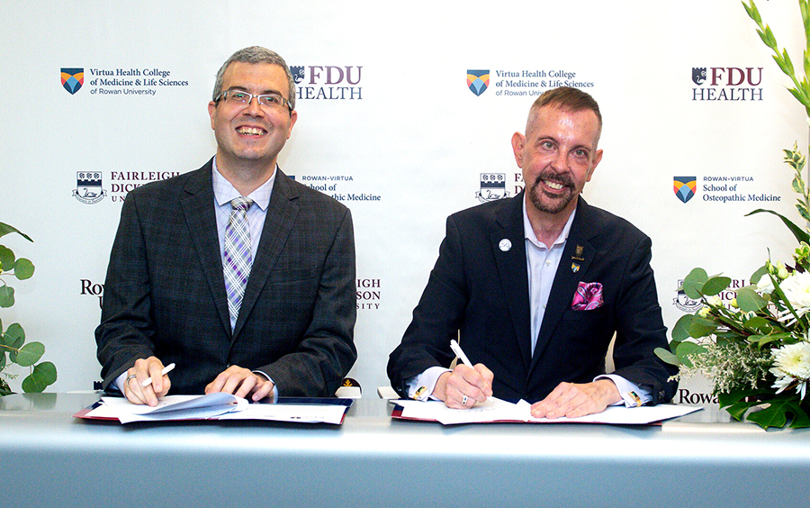 two men sit at a table and sign papers. they smile at the camera.