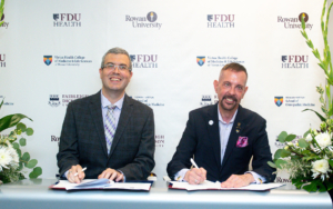 two men sit at a table and sign papers. they smile at the camera.