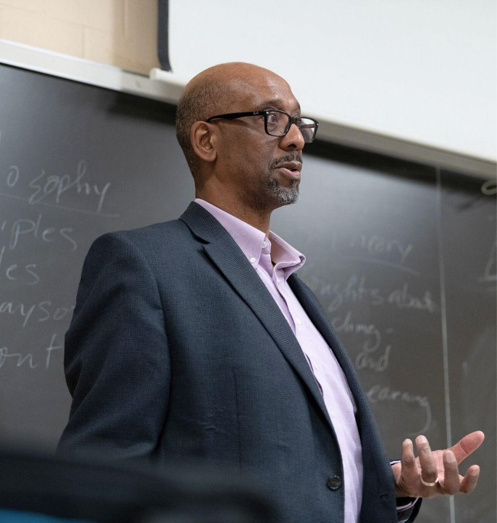 a professor stands at a chalkboard and speaks to a class out of frame.