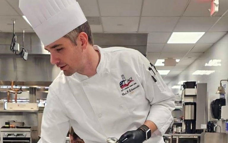 A man wearing a tall chef's hat and chef's coat works in a kitchen.