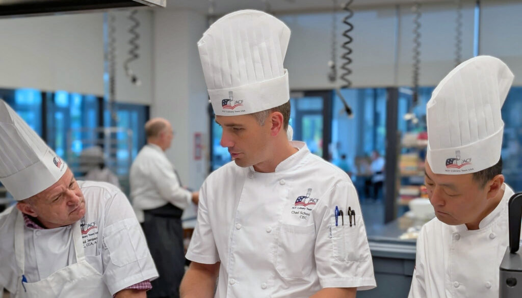 Three men in chefs hats talk in the kitchen.