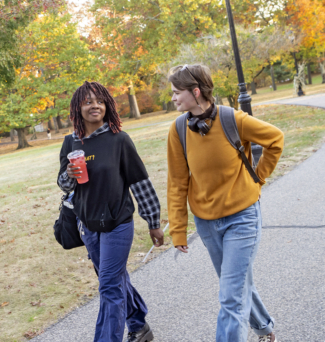 Two students walk on a sidewalk on campus. The leaves on the trees behind them are changing to fall colors.