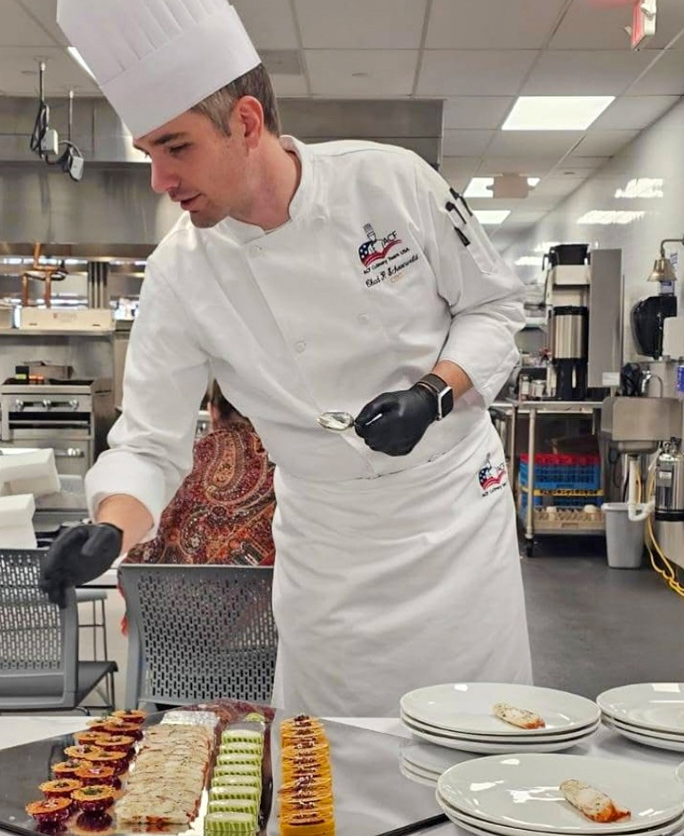 A man wearing a tall chef's hat and chef's coat works in a kitchen.