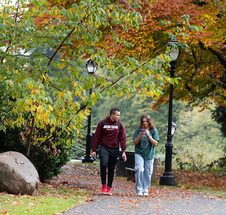 Two students walk down a path at the Florham Campus. The leaves on the trees are turning fall colors.