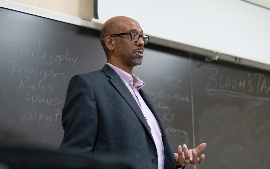 a professor stands at a chalkboard and speaks to a class out of frame.