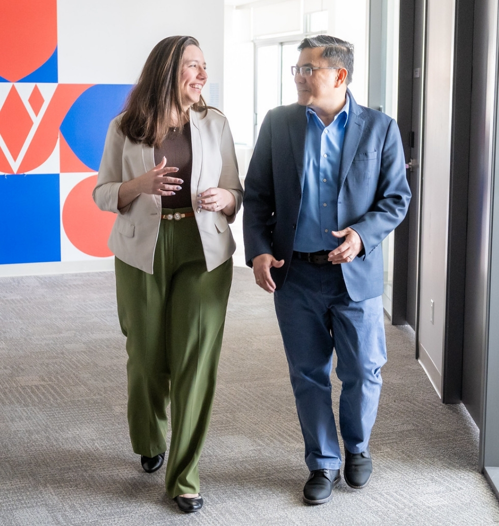 a woman and a man chat as they walk down a hallway. they are dressed in casual business attire.