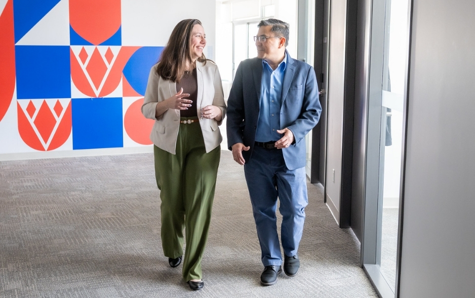 a woman and a man chat as they walk down a hallway. they are dressed in casual business attire.