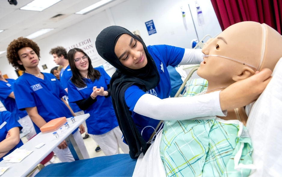a nursing student puts a nasal cannula onto a patient mannequin. other nursing students watch behind her.