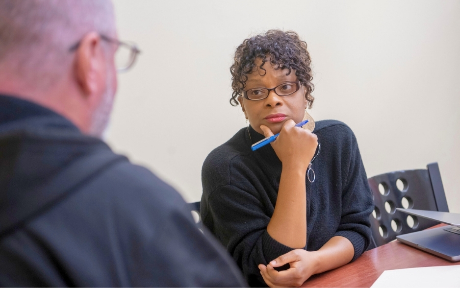 a woman listens to a man. they sit at a table. the woman holds a pen.