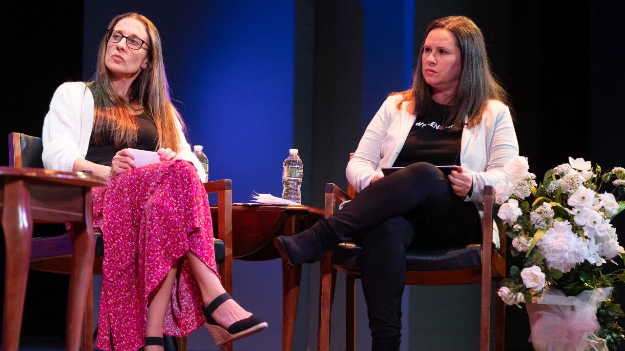 two women sit in chairs and look at someone out of frame.