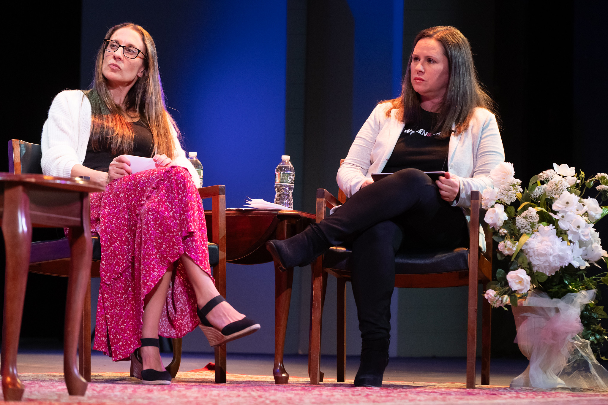 two women sit in chairs and look at someone out of frame.