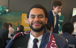 a student wears a suit and holds up a certificate. he is wrapped in a FDU flag.
