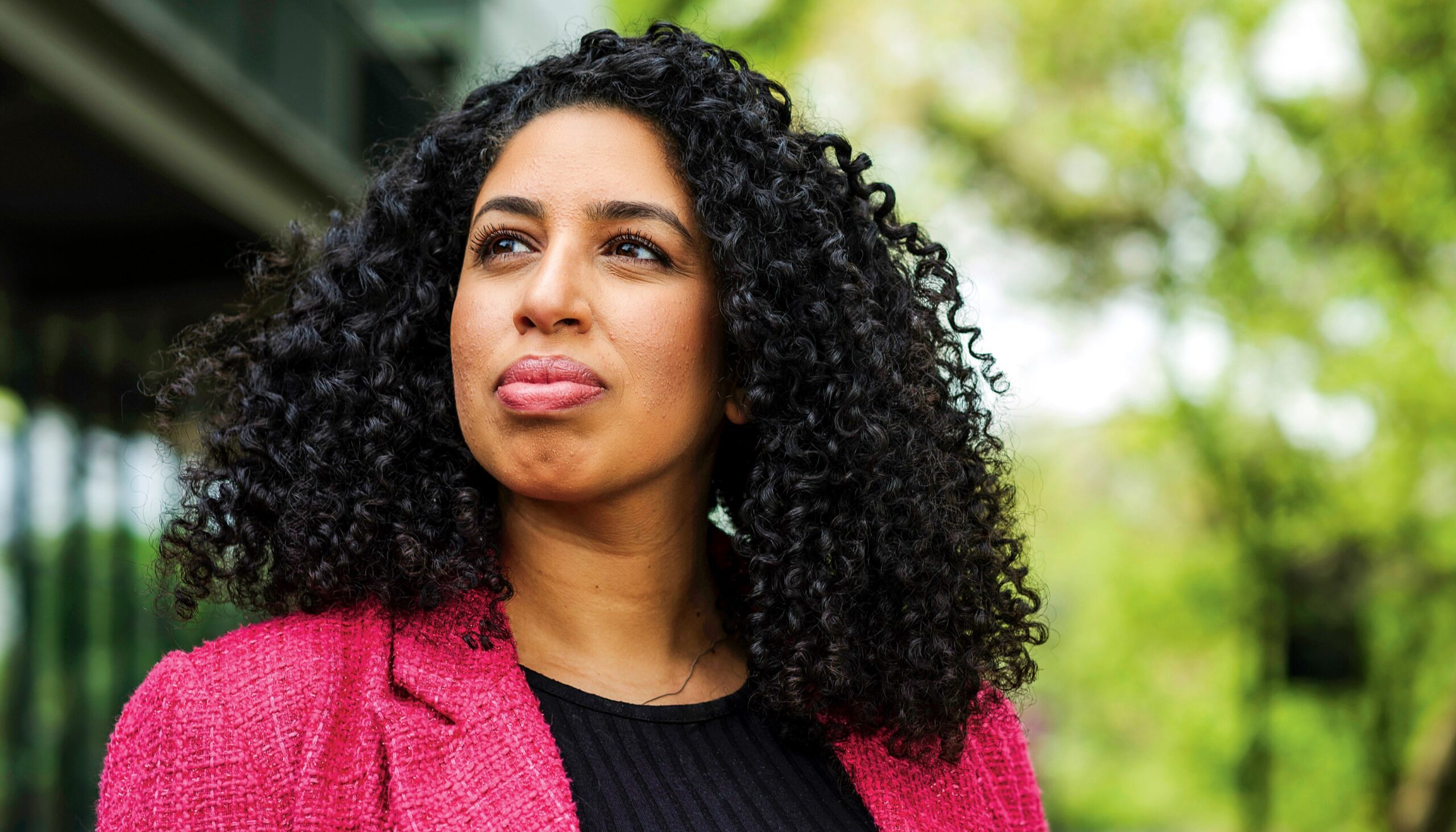 A woman wearing a pink blazer stands outside, gazing off into the distance.