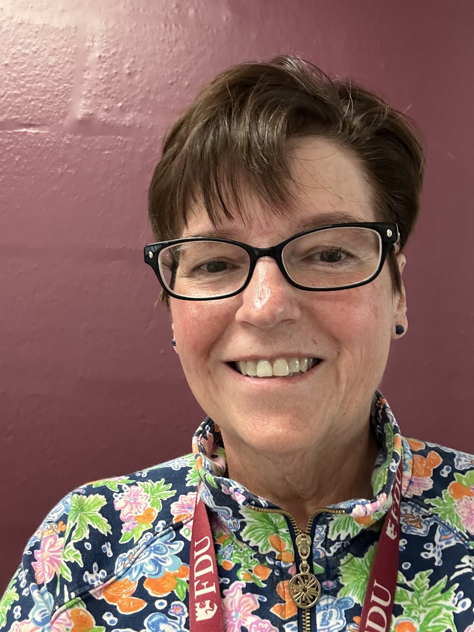 Picture of Terry van Auken infront of a maroon background with a flower patterned jacket on, smiling at the camera.