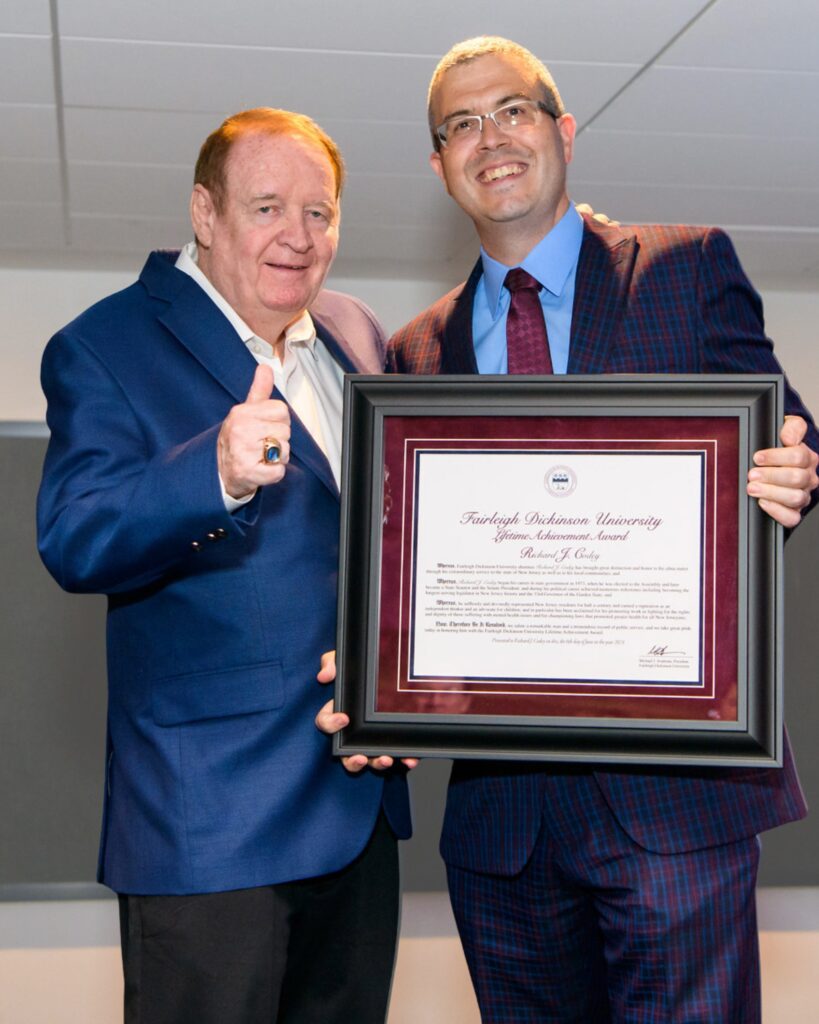Two men pose for a photo with a framed citation. One man gives the thumbs up.