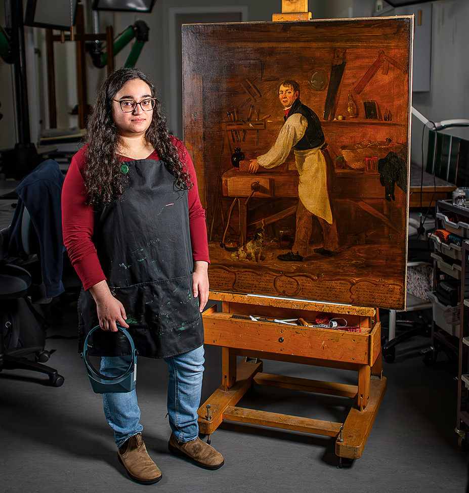 A woman stands next to a canvas painting in an art restoration studio.