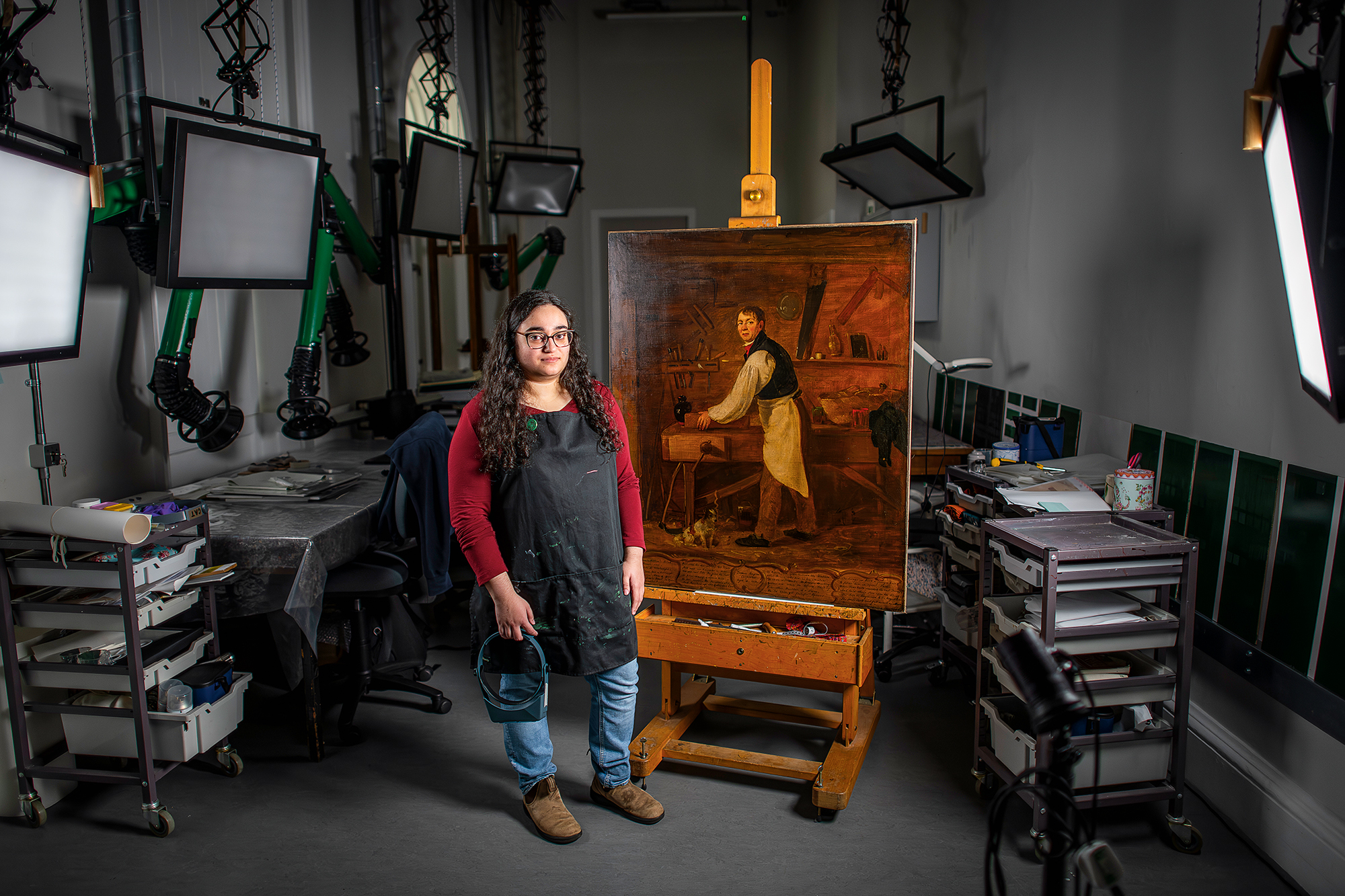 A woman stands next to an easel painting in an art conservation studio.