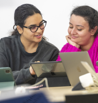 Two female students chat in class while working on their laptops.