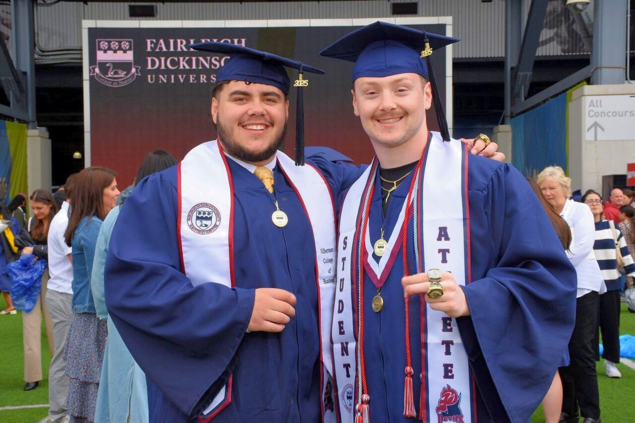 two graduating students smile at the camera. they wear caps and gowns regalia.