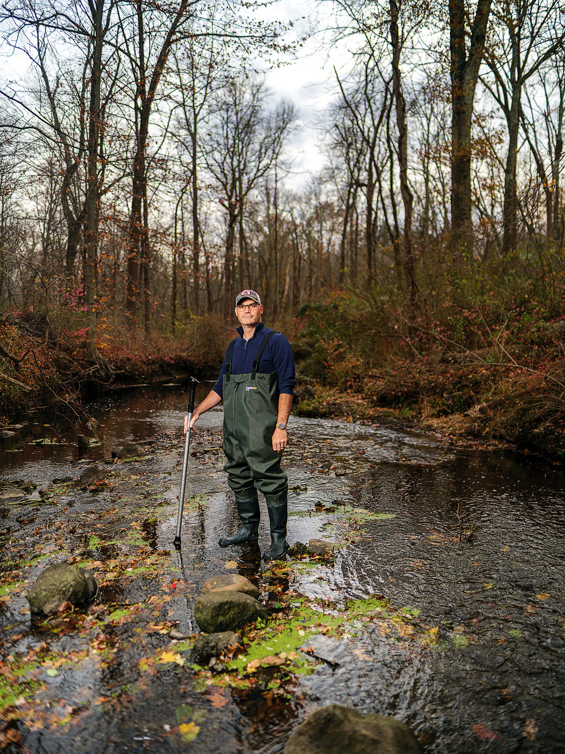 A man stands in the middle of a brook, with running water, surrounded by trees.