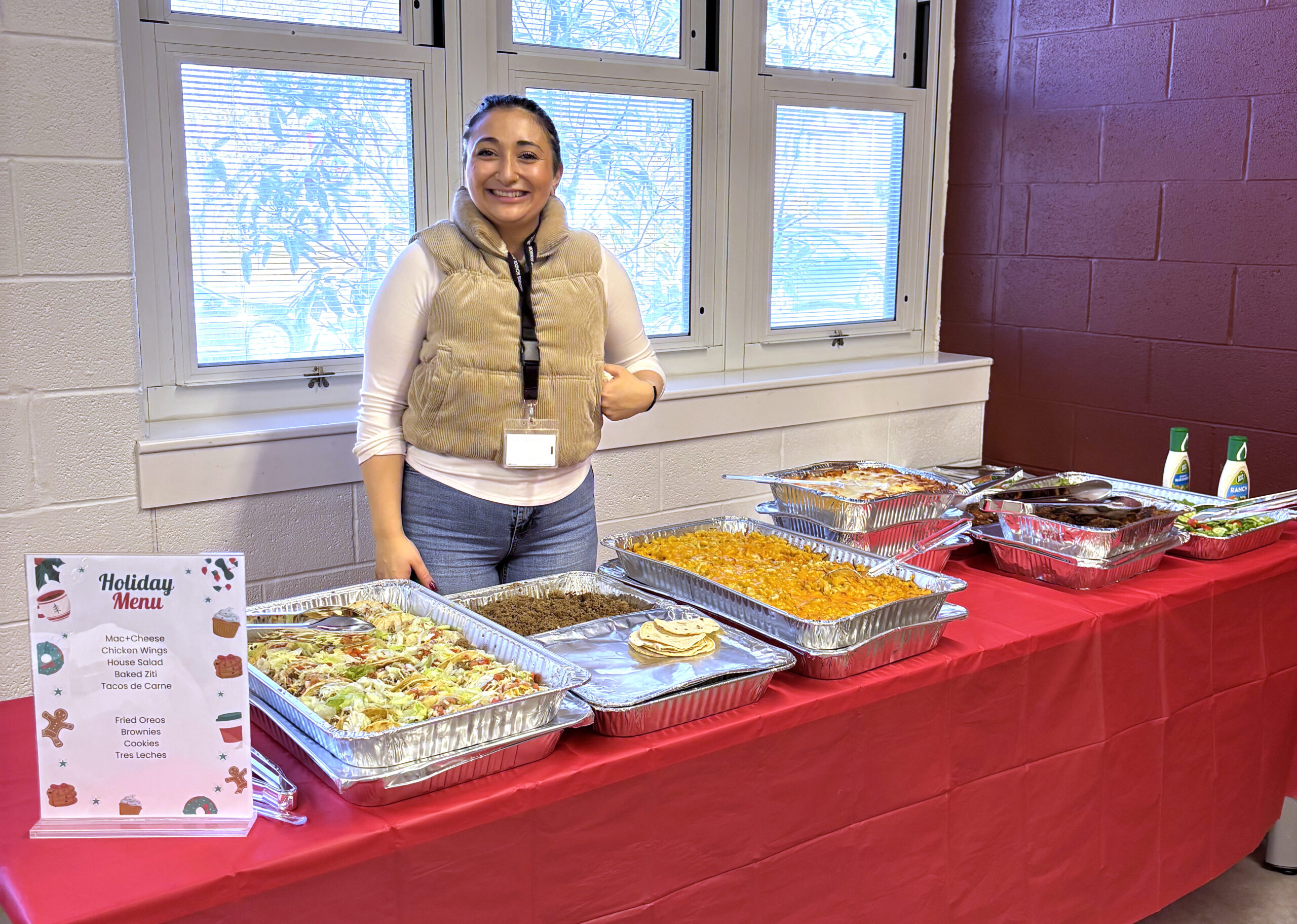 A woman stands in front of windows and behind a table of food.
