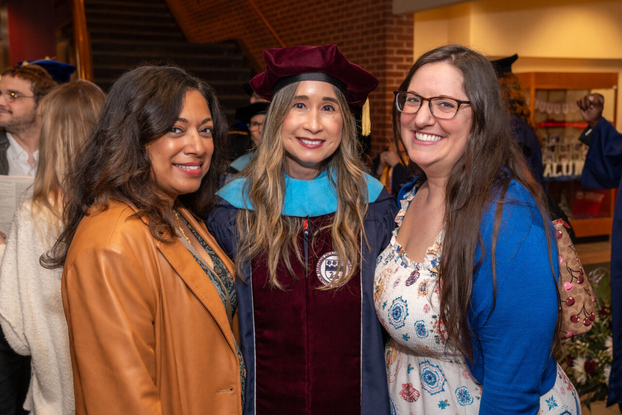three women smile for the camera. one wears regalia.