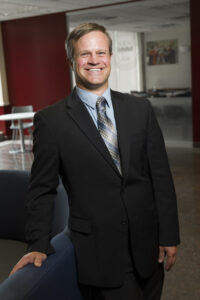 a man wears a suit, stands and smiles for the camera.