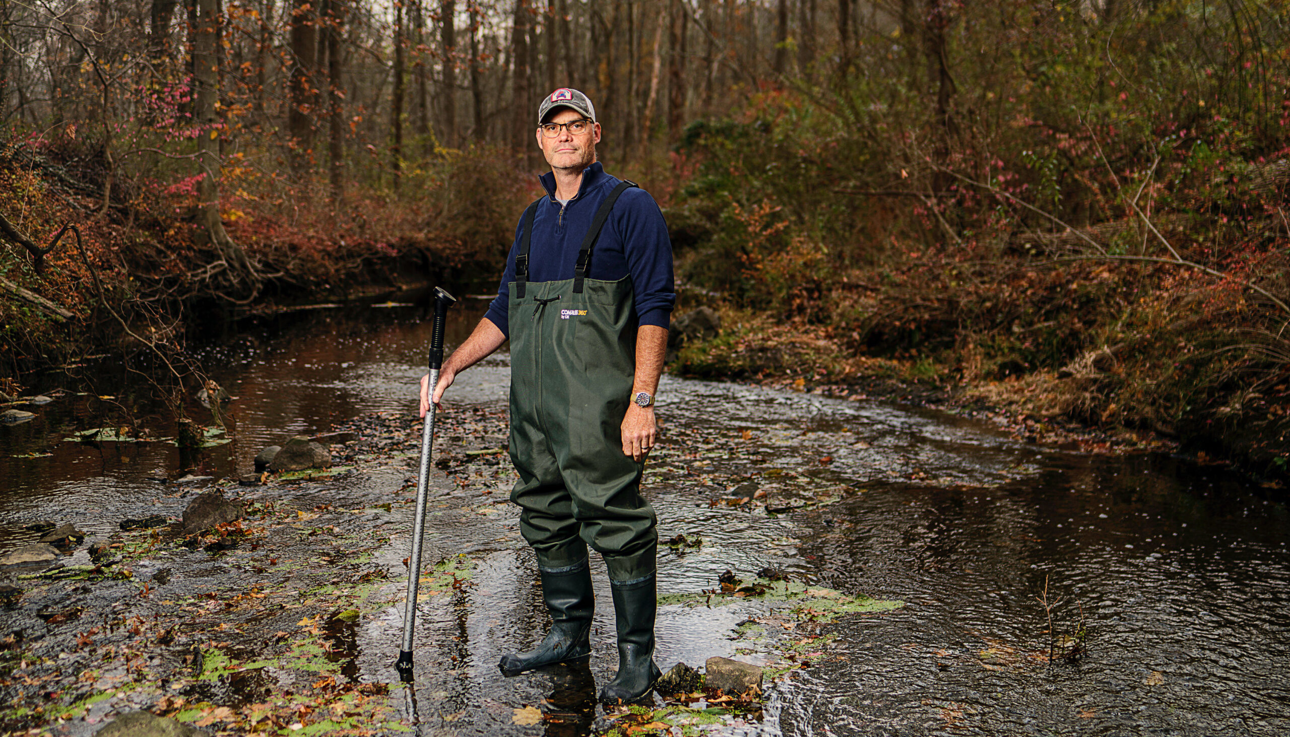 A man stands in the middle of a brook, with running water, surrounded by trees.