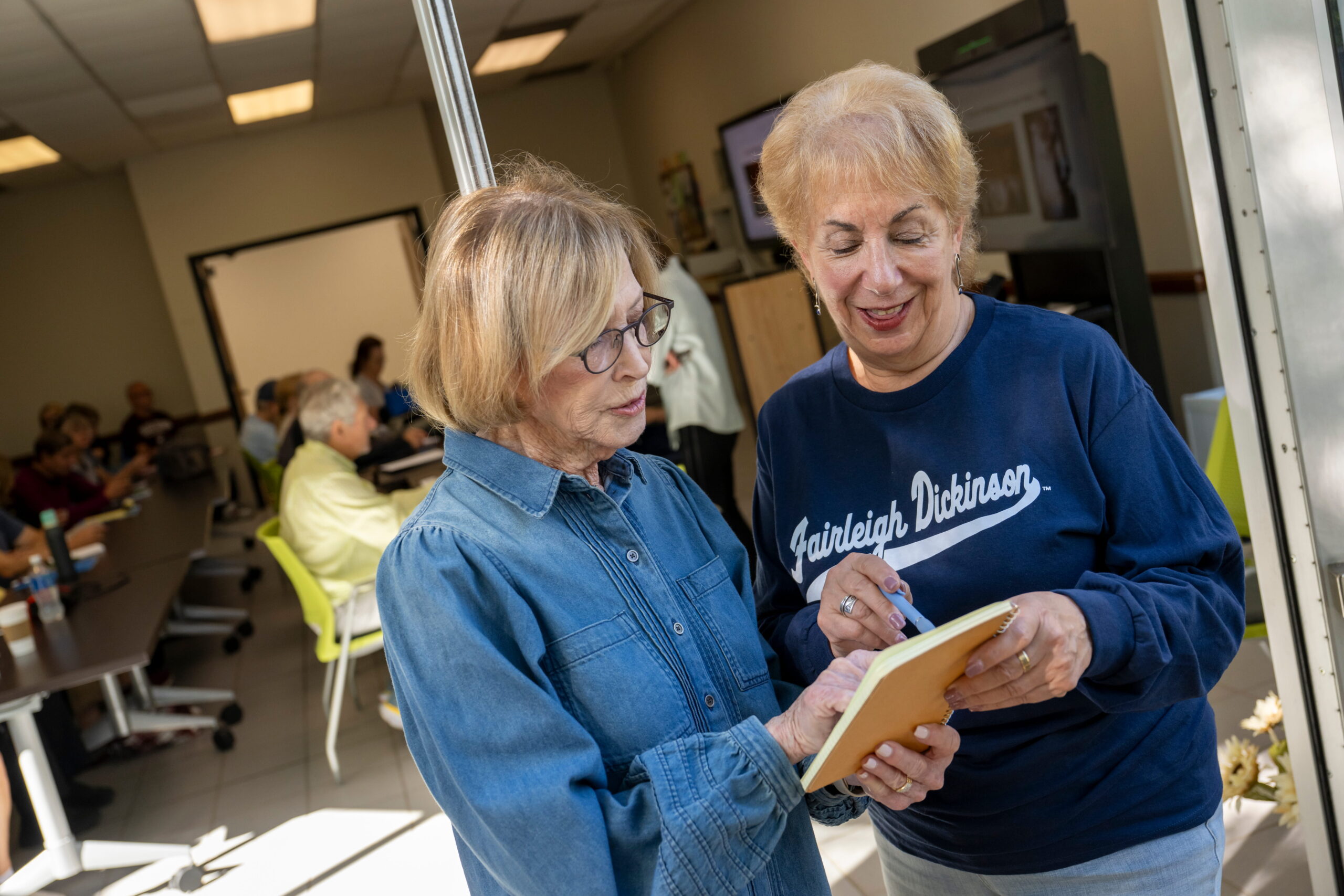 two older women talk together and point at a notepad. they stand outdoors with a classroom behind them.