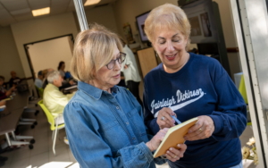 two older women talk together and point at a notepad. they stand outdoors with a classroom behind them.