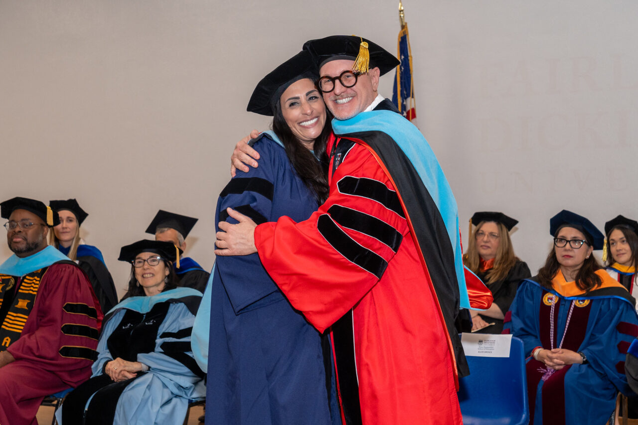 a professor and a graduating student embrace on a stage and smile at the camera. they both wear academic regalia.