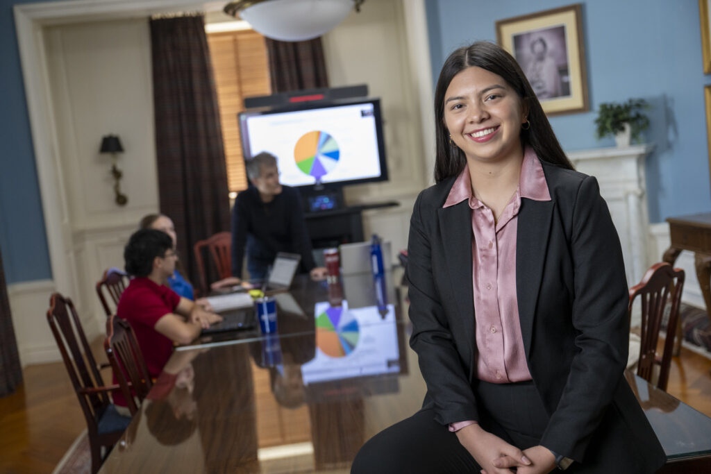 a college student wears a blazer and leans on a table. behind her, there is a presentation with a pie chart.
