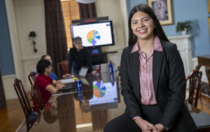 a college student wears a blazer and leans on a table. behind her, there is a presentation with a pie chart.