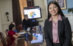 a college student wears a blazer and leans on a table. behind her, there is a presentation with a pie chart.