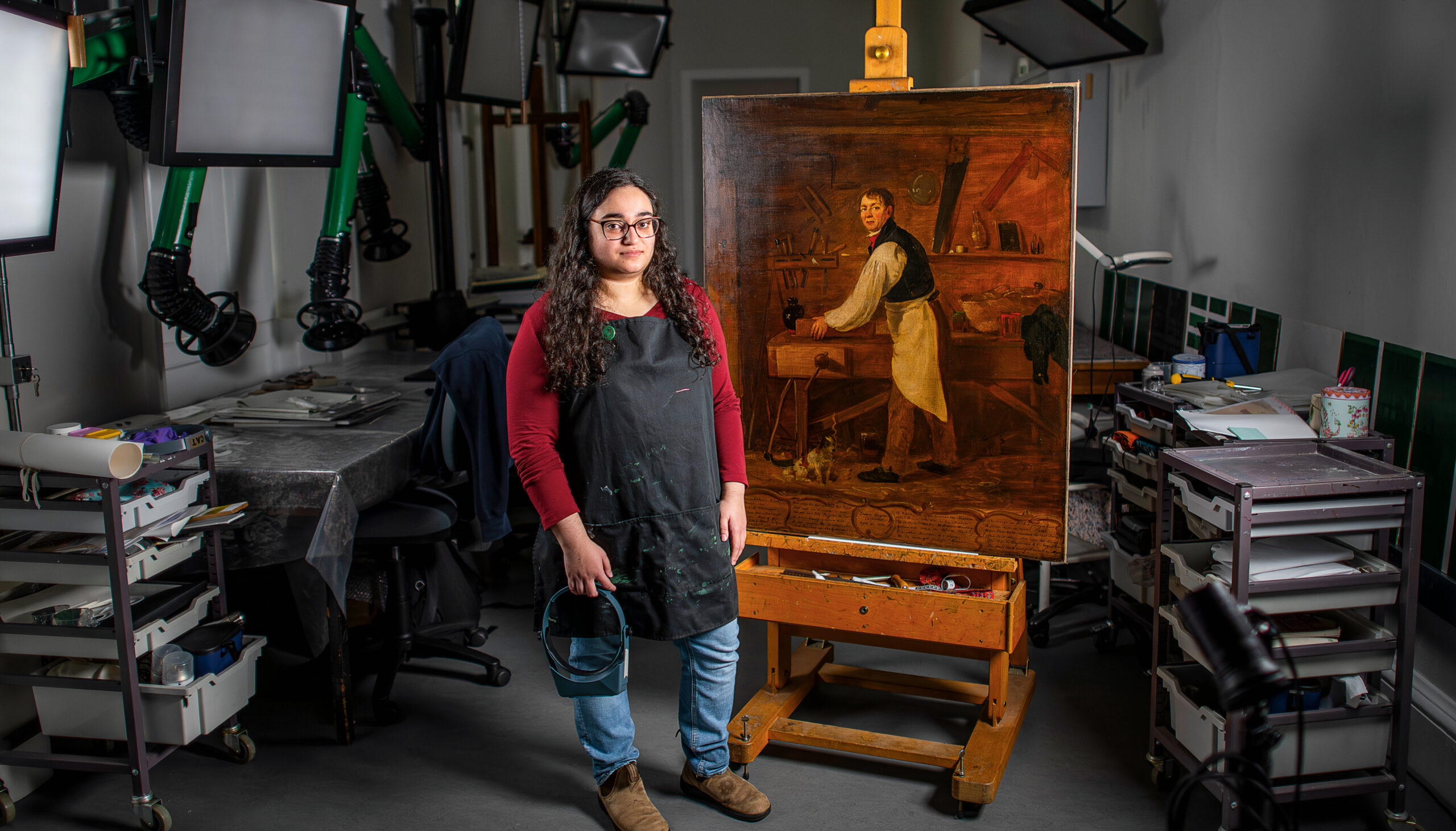 A woman stands next to an easel painting in an art conservation studio.