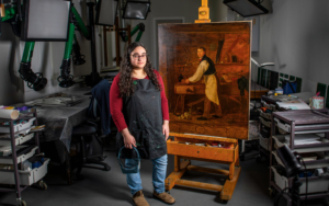 A woman stands next to an easel painting in an art conservation studio.