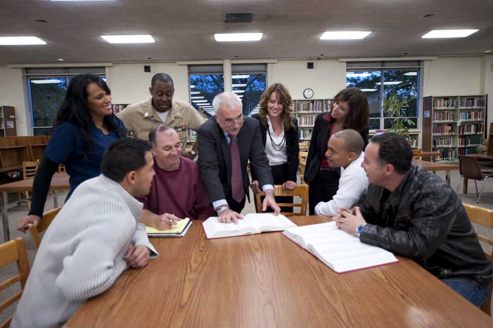 A group of eight adults gathered around a wooden table in a library setting, engaged in a collaborative discussion. An older man with white hair in a dark suit stands at the center, pointing to an open book on the table. The group includes people of various ages and backgrounds, some seated and some standing, all focused on the materials spread across the table which include large open books and notebooks. Bookshelves, large windows, and fluorescent lighting are visible in the background of the well-lit library space.