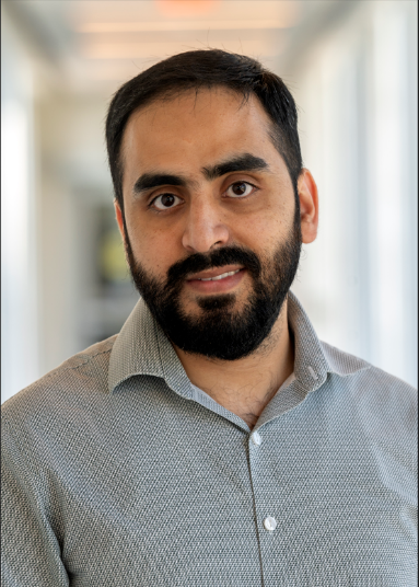 Professional headshot of Dr. Muhammad Umer, a man with short dark hair and a full beard, wearing a light gray patterned button-up shirt, photographed in a softly lit indoor hallway with a blurred background. He is looking directly at the camera with a friendly, approachable expression.