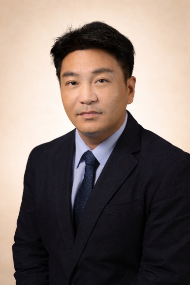 Professional headshot of Dr. Woo Shik Shin, a man with short dark hair, wearing a dark navy suit with a light blue dress shirt and dark patterned tie, photographed against a neutral beige background with soft, professional studio lighting. He has a warm, approachable expression and is looking directly at the camera.