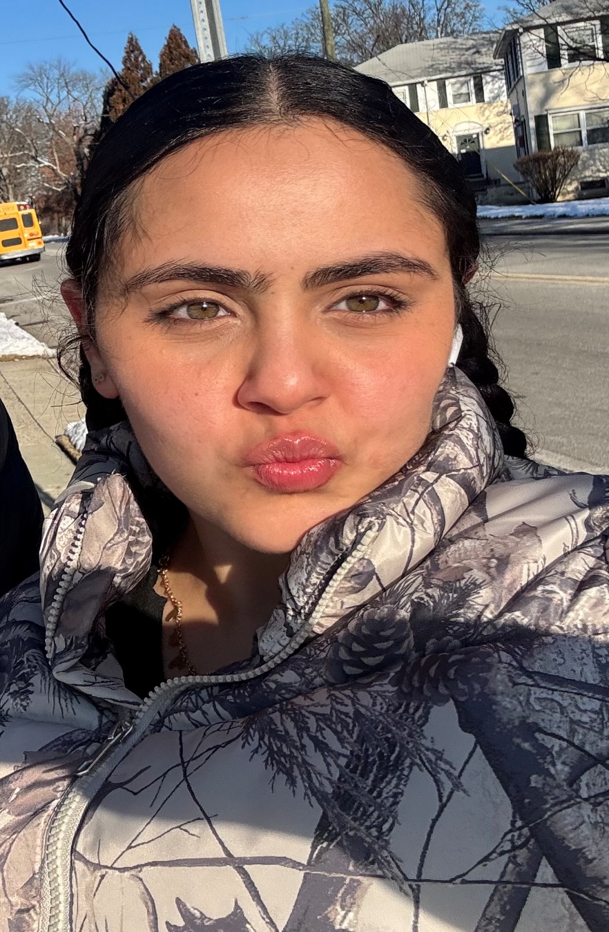 Selfie of a young woman with dark braided hair in a camo jacket outdoors on a sunny winter day, with houses and a school bus visible in the background.