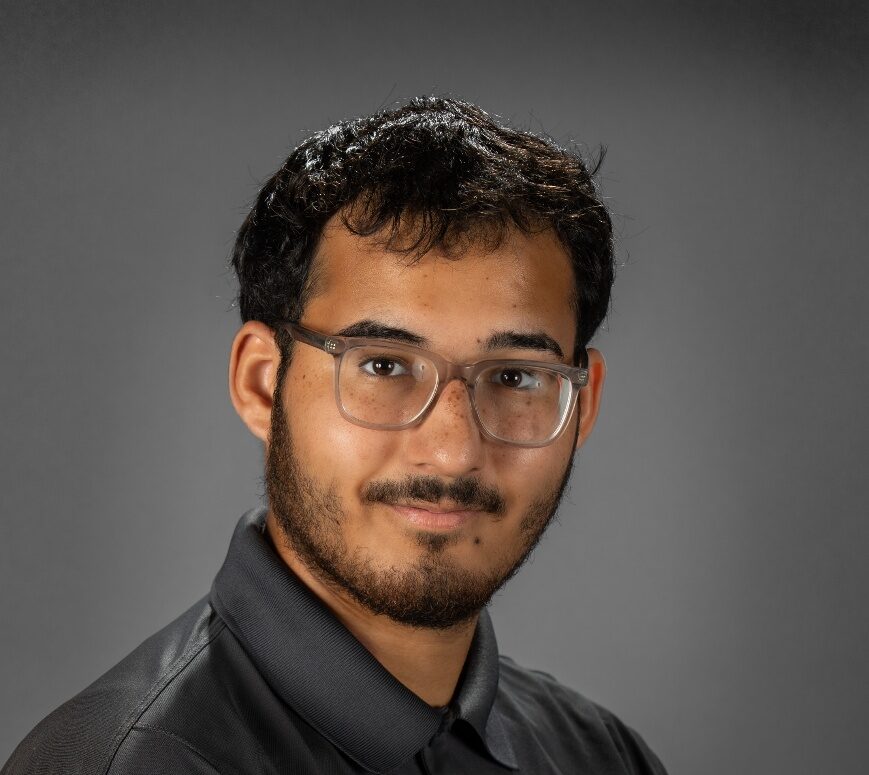 Professional headshot of a young man with short dark hair, a beard, and clear-framed glasses. He is wearing a dark gray polo shirt with the name 'L. CARRION' embroidered in white on the right chest and a white Maltese cross logo containing the number '74' on the left chest. He is smiling slightly against a solid gray studio background.