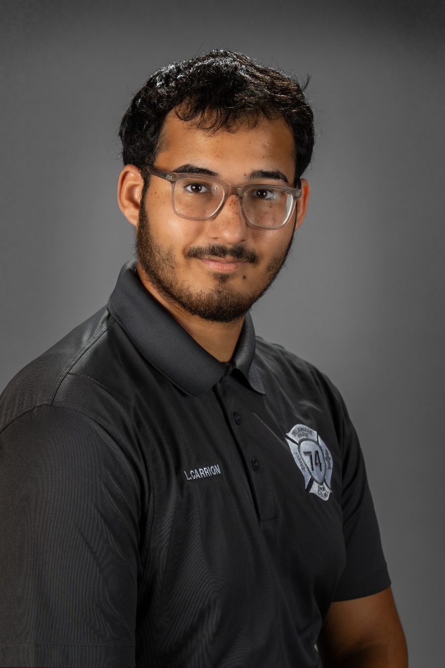 Professional headshot of a young man with short dark hair, a beard, and clear-framed glasses. He is wearing a dark gray polo shirt with the name 'L. CARRION' embroidered in white on the right chest and a white Maltese cross logo containing the number '74' on the left chest. He is smiling slightly against a solid gray studio background.